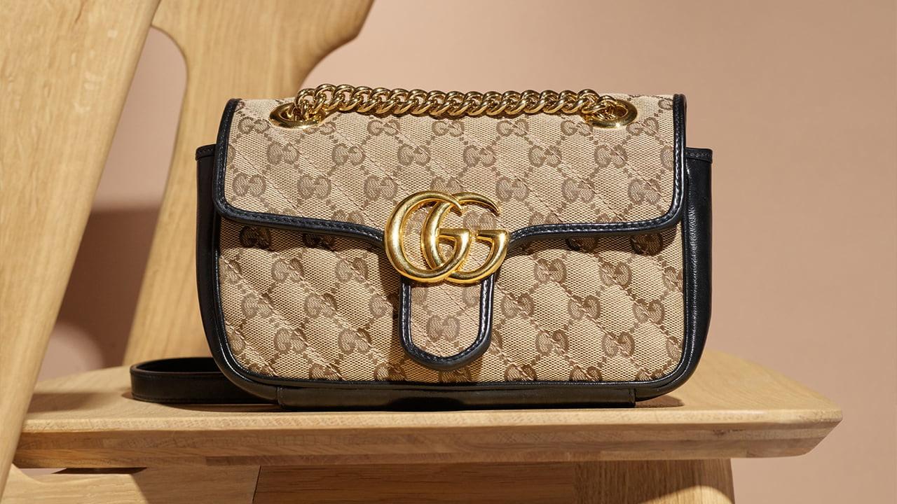 A designer handbag with a monogram pattern, gold chain strap, and a prominent logo on the front, placed on a wooden chair.