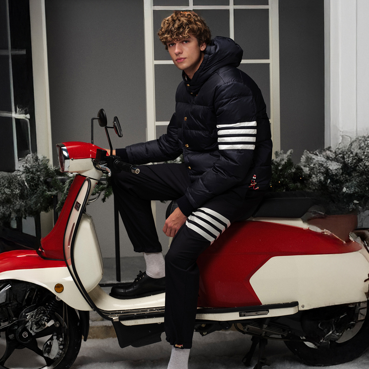 A person in a dark jacket sits on a red and white scooter next to a snowy hedge.