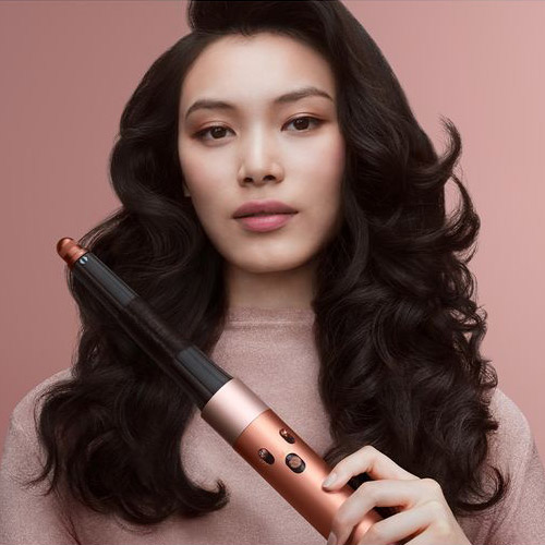 A woman with styled hair holds a Dyson curling tool against a soft pink background.
