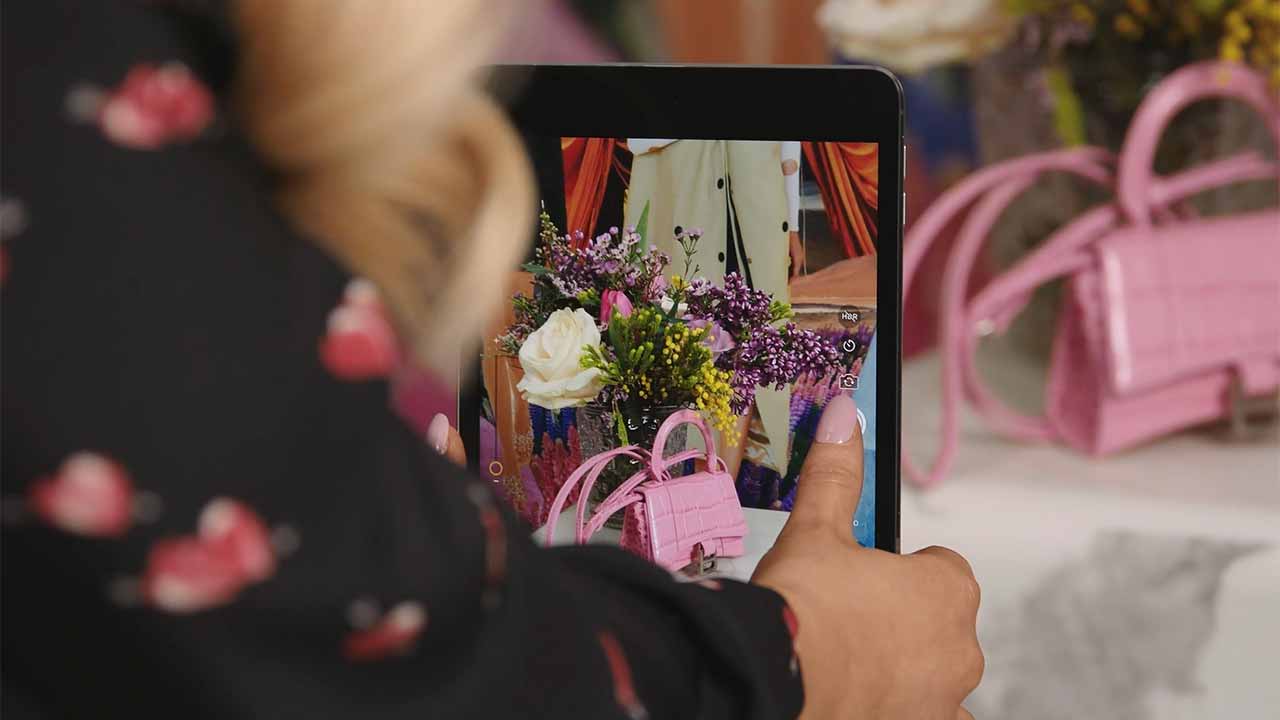 A person photographs purple and white flowers with a tablet; pink handbags are visible in the background.